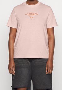 Světle růžové bavlněné tričko s krátkými rukávy, na kterém je v tmavě oranžové barvě vytištěno "Calvin Klein Jeans" a "1978". Ležérní, pohodlný střih.