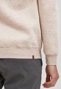 Lysebeige sweatshirt med ribbenede manchetter og kant, forsynet med et lille brunt lædermærke. Bæres med mørkegrå bukser.