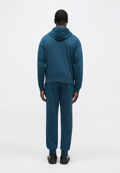 Mätová mikina s kapucňou a rebrovanými manžetami, v kombinácii s rovnakými teplákmi. Oblečenie má voľný strih a mäkkú textúru.