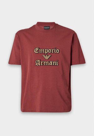 Jeune homme debout, une main dans la poche, portant un t-shirt Emporio Armani bordeaux, un jean large bleu clair et des chaussures noires sur fond blanc.
