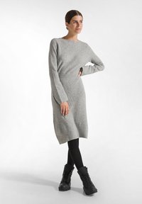 Graues Strickkleid mit langen Ärmeln, rundem Ausschnitt und seitlichen Schlitzen, kombiniert mit schwarzen kniehohen Stiefeln und schwarzen Leggings.