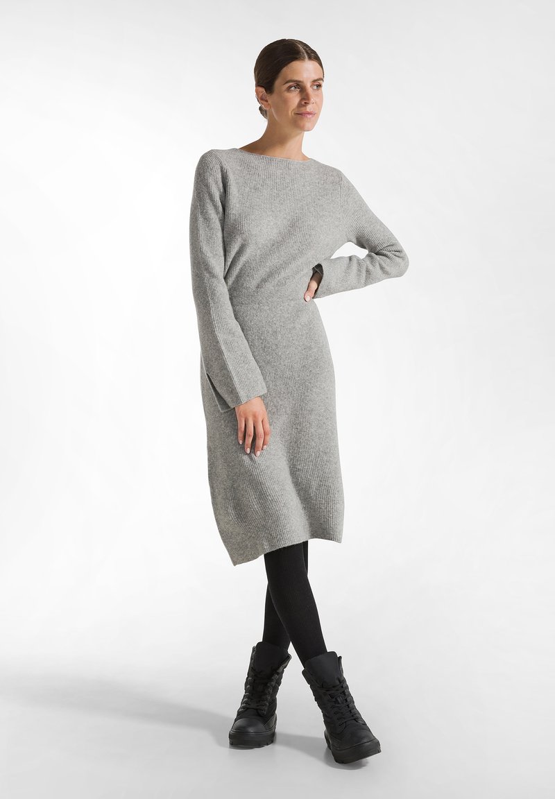 Graues Strickkleid mit langen Ärmeln, rundem Ausschnitt und seitlichen Schlitzen, kombiniert mit schwarzen kniehohen Stiefeln und schwarzen Leggings.