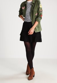 Femme portant une veste ornée de patchs verts, une jupe noire à volants, des collants noirs, des bottines brunes et un haut à rayures noir et blanc.