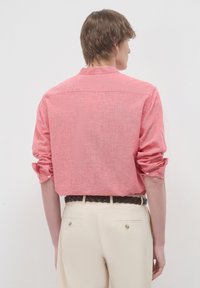 Camicia rossa con texture, maniche arrotolate, polsini abbottonati e un design con yoke sul retro, abbinata a pantaloni beige e una cintura intrecciata.