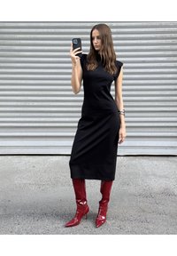 Czarna dopasowana sukienka z krótkimi, strukturalnymi rękawami, do kolan. Noszona z błyszczącymi, czerwonymi, wysokimi na stiletto kozakami do kolan. Modelka trzyma smartfona.