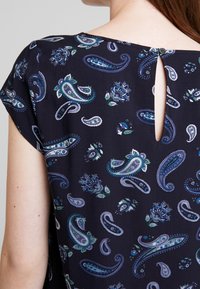 Blusa azul marino oscuro con patrones de paisley y flores. Cierre en la parte posterior con forma de ojo de cerradura, mangas cortas, tela ligera con una textura suave.