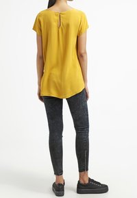 Haut jaune à manches courtes avec un dos à découpe en forme de clé, associé à un jean skinny en denim foncé présentant une texture délavée et des zips aux chevilles. Des baskets noires complètent le look.