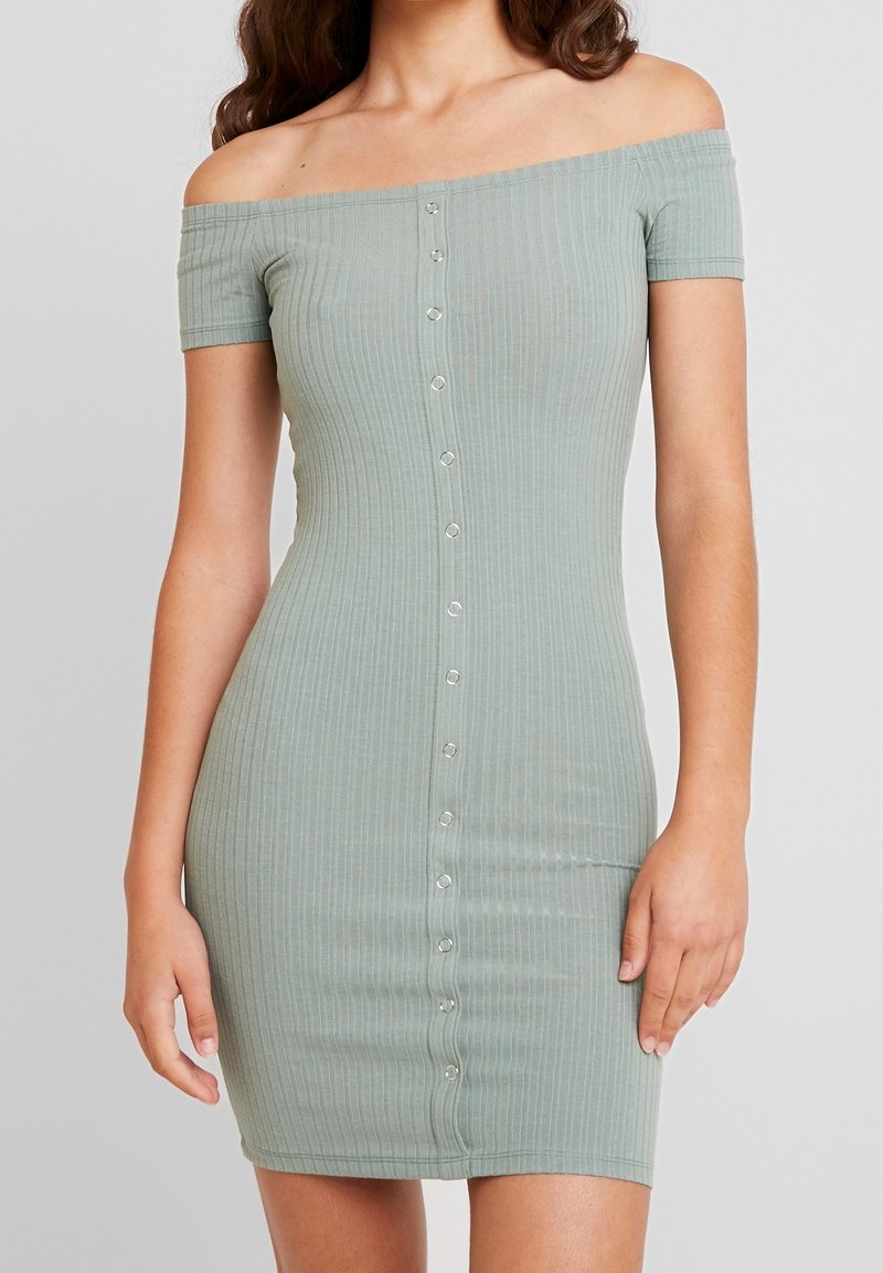 Femme portant une robe ajustée, à manches courtes et épaules dénudées en maille côtelée vert clair, avec une rangée de boutons au centre devant.