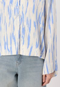 Locker sitzende, knopffrontierte Bluse in Creme mit blauem abstraktem Pinselstrichmuster. Verfügt über lange Ärmel und einen abgerundeten Saum. Kombiniert mit hellblauem Denim.
