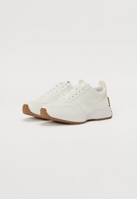 Białe sportowe buty z gładką cholewką, teksturowaną gumową podeszwą i kontrastującą beżową podeszwą. Posiada okrągłe sznurowadła i minimalistyczny design.