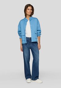 Light blue, textured jacket with a stand collar, front pockets, and buttons. Paired with dark blue wide-leg jeans and white sneakers.