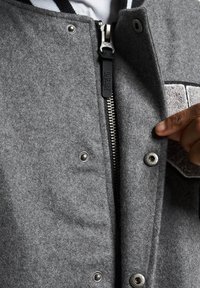 Grå ullblanding jakke med frontglidelås, trykknapper og en teksturert lapp. Stripete fôr synlig ved kragen.