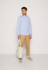 Lichtblauw overhemd met knopen, beige chino's, suède schoenen, met een crèmekleurige totebag. Eenvoudig ontwerp, overhemd met kraag en lange mouwen, casual look.