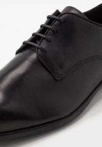 Chaussure de ville en cuir noir avec une surface lisse, bout arrondi, cinq œillets pour les lacets et des détails de couture subtils le long du bord.