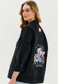 Czarna kurtka dżinsowa z kolorowym nadrukiem w kształcie kokardy z tyłu, z napisem "Każdy odcień przykrywa kształt" i "Wszyscy jesteśmy BOW-TIFUL." Szczegół z czerwoną łatą.
