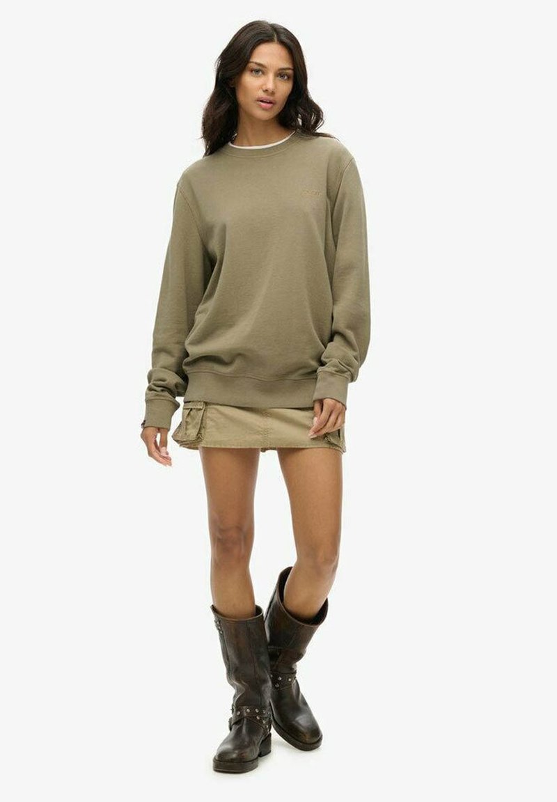 Sudadera color caqui con cuello redondo, mangas largas y puños acanalados, combinada con una falda beige y botas marrones hasta la rodilla con tachuelas.