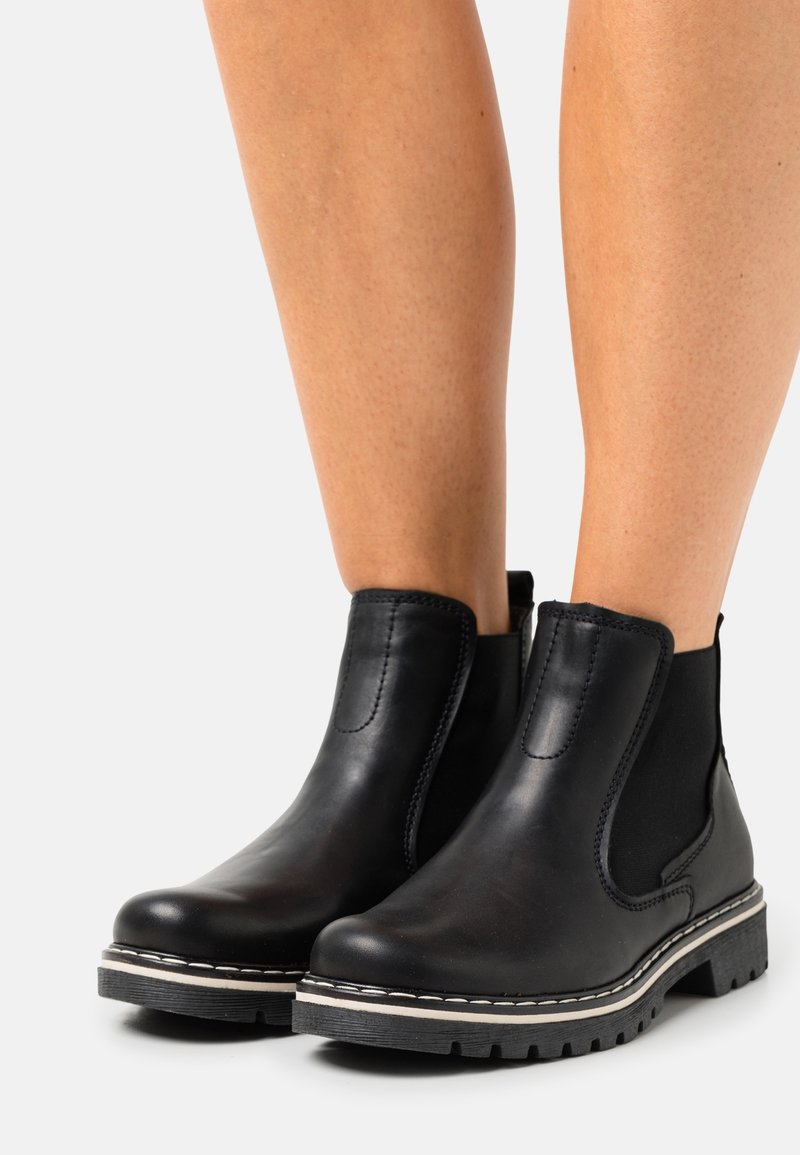 Personne portant des bottines Chelsea en cuir noir avec panneaux latéraux élastiques et semelles en caoutchouc épaisses, debout sur un fond blanc.