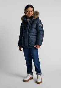 Marinblå pufferjacka med foder i päls, dragkedjestängning och sidofickor. Bärs med blå jeans och vita sneakers med bruna sulor.