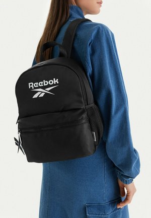 Mujer con vestido azul lleva una mochila Reebok negra con bolsillo delantero con cremallera y logo en la parte trasera.