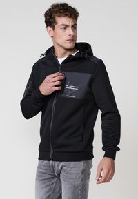 Sudadera con cremallera negra con textura acanalada, un bolsillo frontal en contraste y cordones en la capucha. Presenta un diseño elegante y puños ajustados.