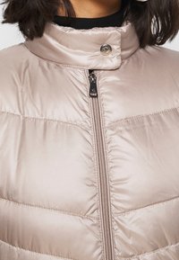 Prošívaná vesta ve světlé béžové barvě, s vysokým límcem, předním zipem s kovovým taháčkem a prošívanou texturou s prošitými detaily.