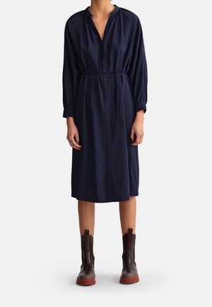 Vrouw draagt een marineblauwe midi-jurk met knopen, een tailleband en lange mouwen, gecombineerd met donkerbruine enkellaarzen met dikke zolen.