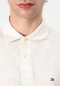 Slonobiely polo tričko s jemným vzorom listov, s tromi gombíkmi a malým logom na ľavej strane. Bavlnený materiál, krátke rukávy.
