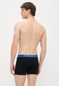 Μαύρα ανδρικά εσώρουχα μπόξερ με μπλε μέση που φέρει το σήμα "BOSS". Το υλικό φαίνεται λείο, εφαρμοστό με κλασικό σχεδιασμό.