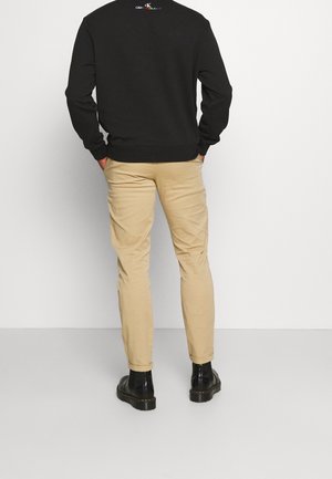 Mies pukeutuneena mustaan Calvin Kleinin puseroon, beigeihin kapeisiin housuihin ja mustiin nahkasaappaisiin, seisoo valkoista seinää vasten, kädet taskuissa.