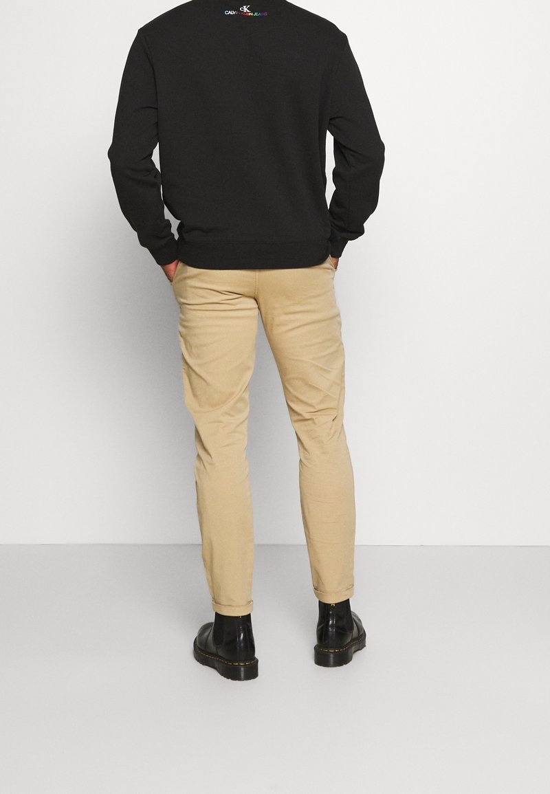 Homme portant un sweat-shirt noir Calvin Klein, un pantalon beige slim-fit et des bottes en cuir noir, debout contre un mur blanc, les mains dans les poches.