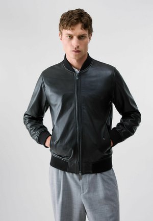 Giovane uomo con capelli corti e ricci, indossa una giacca di pelle nera e pantaloni grigio chiaro, in piedi davanti a uno sfondo chiaro e uniforme, con le mani nelle tasche.