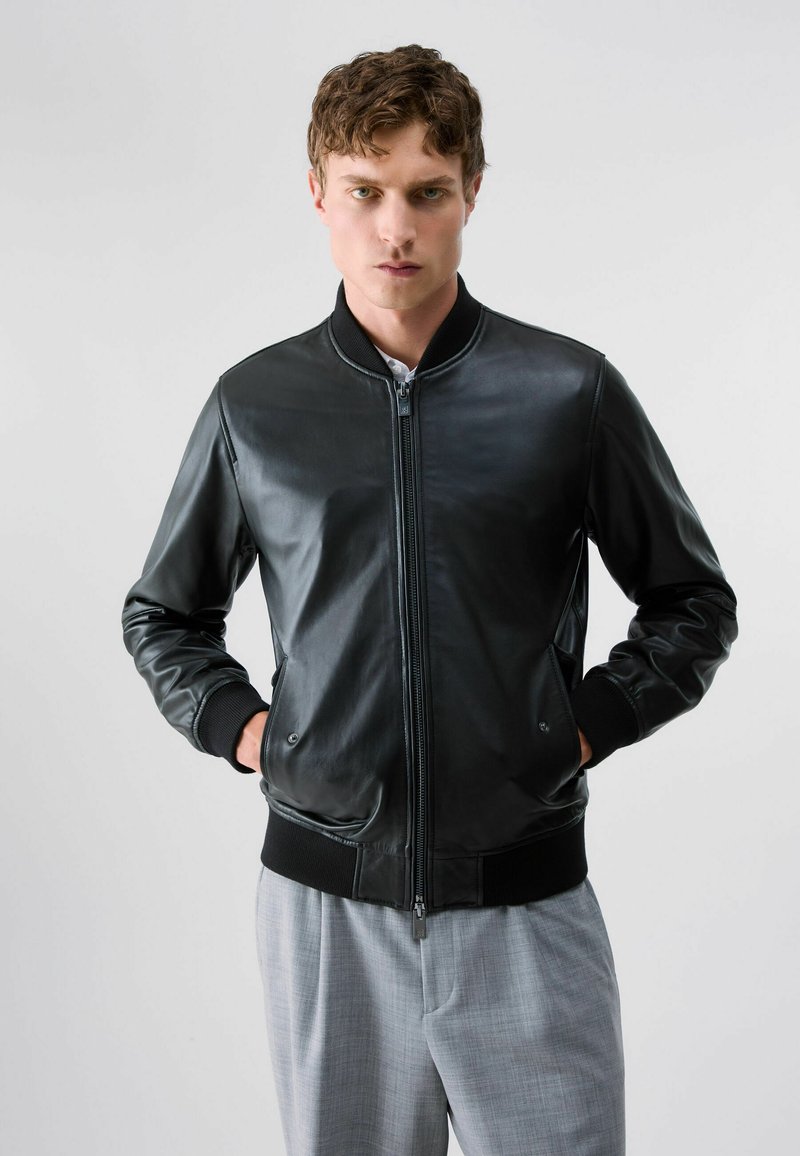 Jeune homme aux cheveux courts et bouclés portant une veste en cuir noire et un pantalon gris clair, debout devant un fond clair uni, les mains dans les poches.