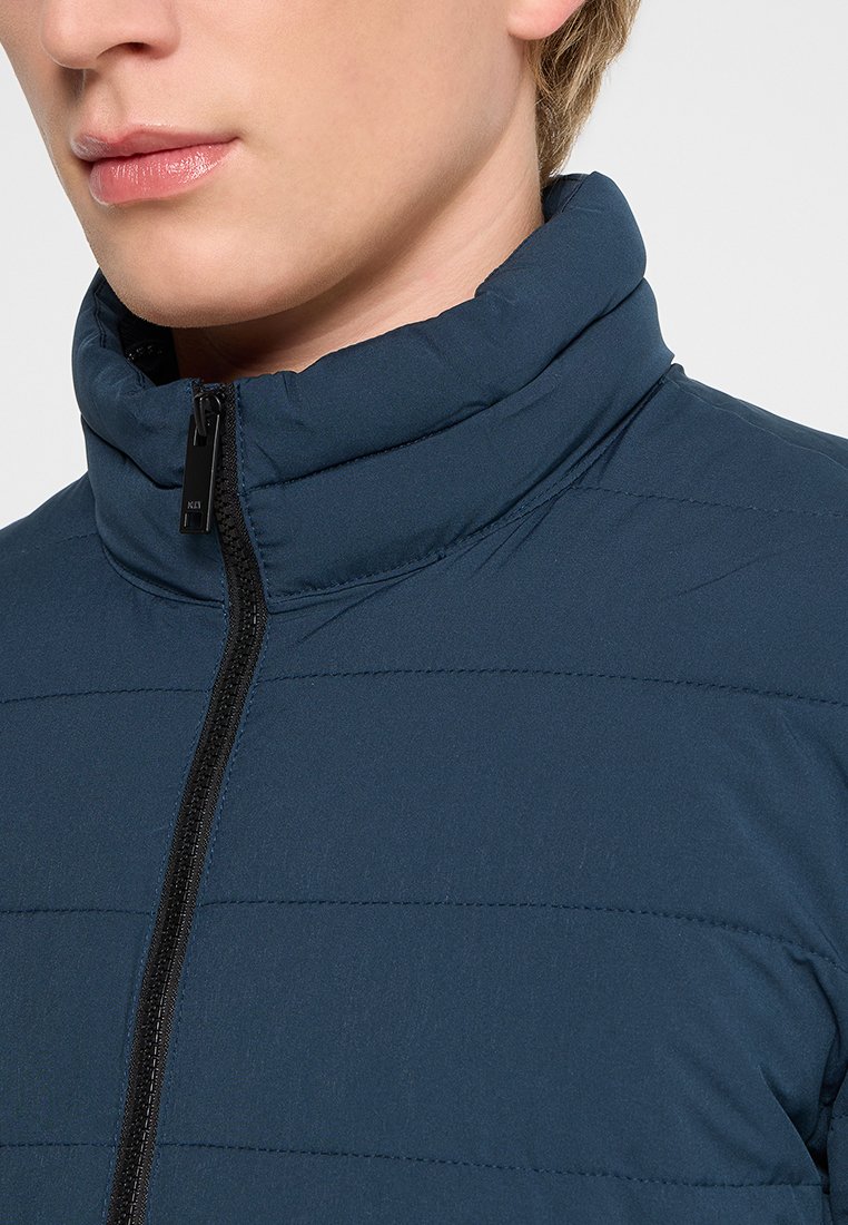 Marineblauwe gewatteerde jas met hoge kraag, ritssluiting aan de voorkant en horizontaal gestikt patroon voor textuur en isolatie.