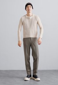 Sweater sem mangas de malha bege sobre uma blusa de manga longa, combinado com calças escuras de oliveira ajustadas e sapatoschunky em duas cores. Gorro preto.