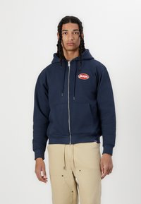 Mörkblå dragkedja hoodie i bomullsblandning, med en kanguroficka fram och röd broderad "HACKER"-logga på bröstet.