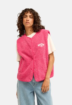 Femme aux cheveux bouclés portant une veste polaire rose vif par-dessus un t-shirt blanc à manches courtes et un jean bleu clair sur un fond uni.