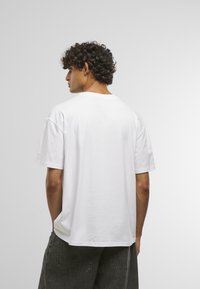 Hvid kortærmet t-shirt, afslappet pasform, rund halsudskæring, bomuldsstof, synlige syninger på skuldrene, båret med løse grå bukser.
