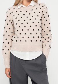 Maglione lavorato a maglia di un rosa chiaro con pois neri pelosi, orlo e polsini a coste, indossato sopra una camicia bianca con colletto, abbinato a pantaloni grigi a pieghe.
