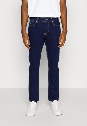 Mand iført mørkeblå straight-leg jeans, hvide sneakers og en hvid skjorte, stående mod en ensfarvet lys baggrund.