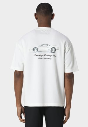 Mężczyzna w białej koszulce z ilustracją samochodu widzianego z boku i napisem „Sunday Racing Club Motor Oil Company” na plecach.
