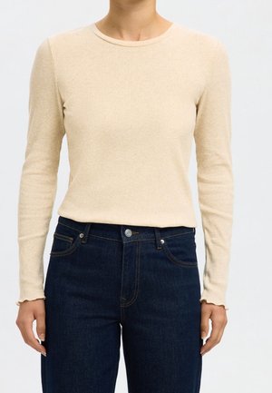 Haut côtelé beige à manches longues associé à un jean bleu foncé taille haute sur une personne vue du cou aux hanches.