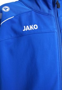 Blaues Sportjackett mit glatter Textur, Reißverschluss und weißem "JAKO"-Logo auf der linken Seite. Akzentnähte an den Schultern.