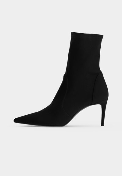 Bottine noire à bout pointu et talon haut et fin, présentée de profil sur fond clair. Design épuré et minimaliste.