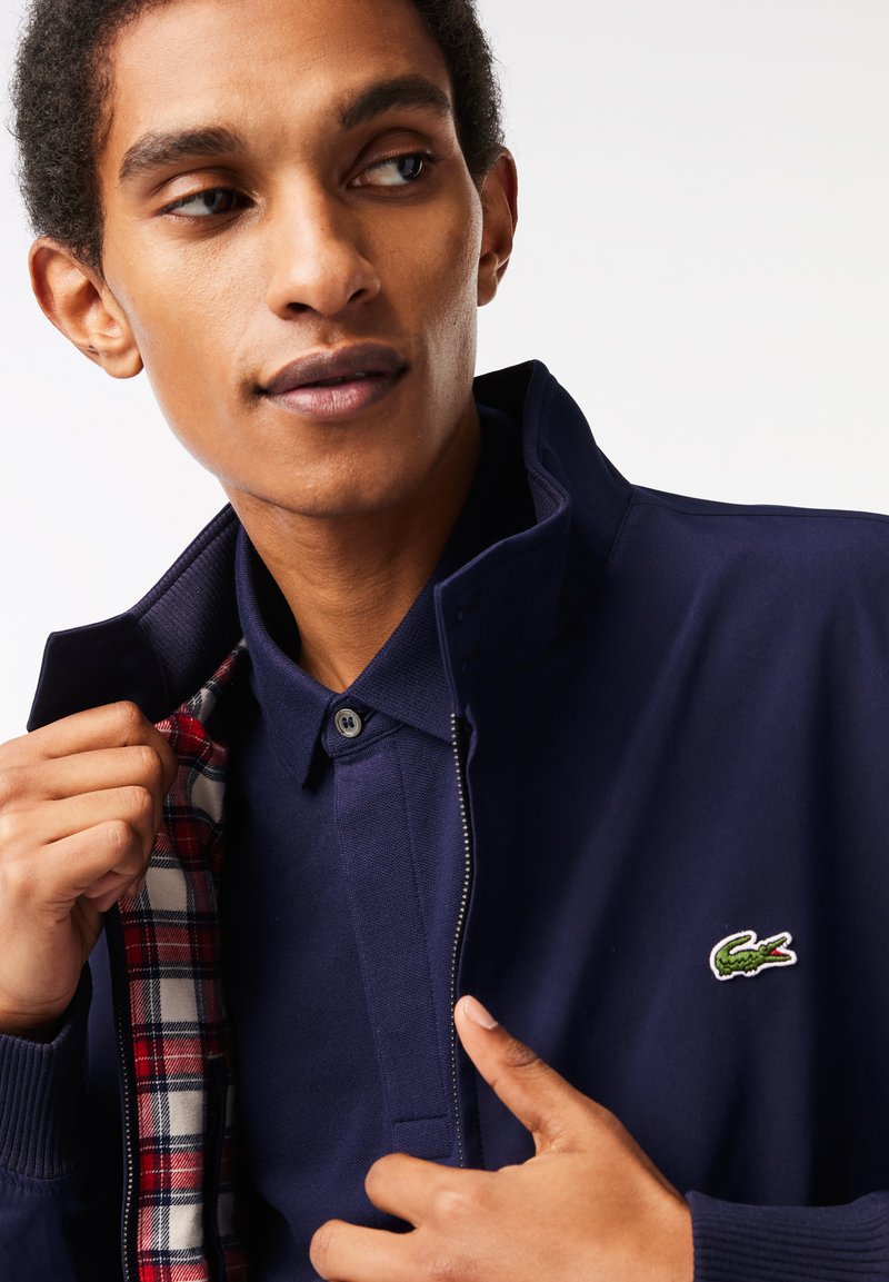 Homme ajustant une veste marine avec une doublure à carreaux, portant une chemise marine boutonnée assortie, avec un logo de crocodile vert sur la poitrine de la veste.