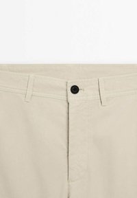 Taille d'un pantalon beige avec un bouton noir, des passants de ceinture et des coutures détaillées au niveau de la braguette à glissière.