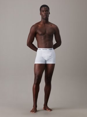 Weiße Baumwoll-Boxershorts mit einem schwarzen elastischen Bund, auf dem das "Calvin Klein"-Logo prangt, getragen von einem männlichen Model mit baren Füßen.
