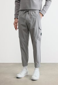 Άνδρας που φοράει ανοιχτό γκρι παντελόνια τύπου cargo jogger με κορδόνι στη μέση, συνδυασμένα με λευκά αθλητικά παπούτσια και λευκές κάλτσες, σε απλό φόντο.