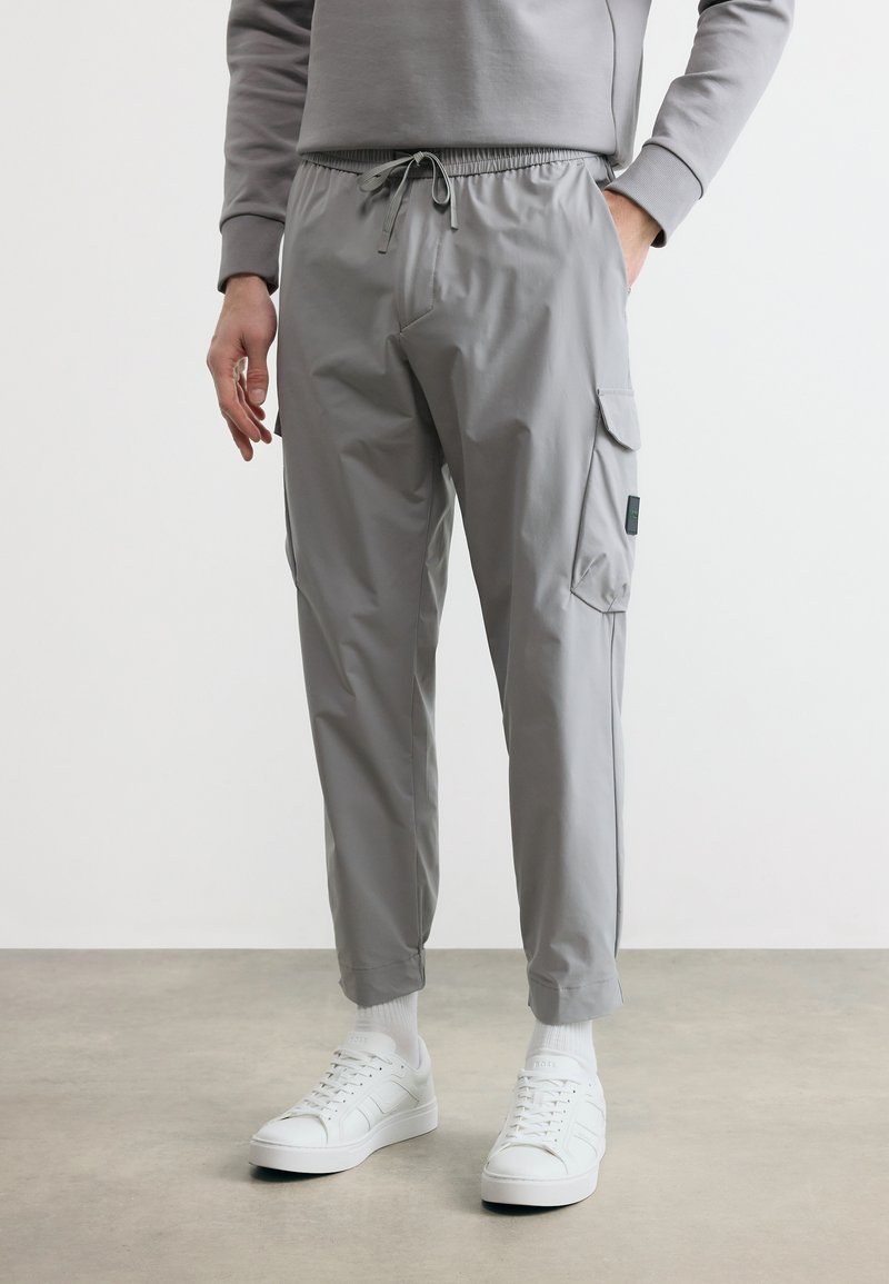 Άνδρας που φοράει ανοιχτό γκρι παντελόνια τύπου cargo jogger με κορδόνι στη μέση, συνδυασμένα με λευκά αθλητικά παπούτσια και λευκές κάλτσες, σε απλό φόντο.