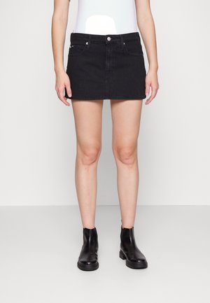 Kvinde iført sort denim mininederdel, hvid top og sorte ankelstøvler, der står mod en ensfarvet lys baggrund.