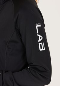 Zwarte sportjas van gladde stof, met een wit 'ELITE LAB' logo op de arm. Beschikt over een aansluitend ontwerp en gestructureerde naden.